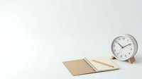 Minimalist workspace with open kraft notebook, wooden pencil, and silver alarm clock on white background, ready for design projects.