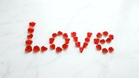 Word Love spelled with red rose petals on white marble surface, romantic Valentine's Day concept for cards and decorations.