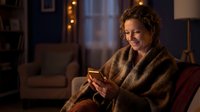 Middle-aged woman with curly brown hair and fair complexion, wrapped in a cozy knit blanket, smiling while holding a warm beverage in a comfortable home setting with warm lighting.