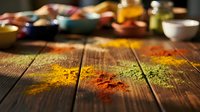 Colorful spices scattered on rustic wooden table with blurred cooking ingredients in background creating warm kitchen atmosphere for culinary projects.