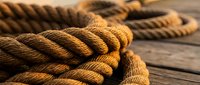 Thick brown nautical rope with detailed twisted fibers coiled on weathered wooden deck surface in warm golden sunlight for marine and sailing themes.