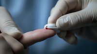 Medical professional performing blood glucose test with lancet device on patient finger wearing protective gloves in clinical setting for diabetes monitoring.