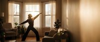 Young woman practicing yoga warrior pose in sunlit living room with warm golden light streaming through windows creating peaceful home wellness atmosphere.