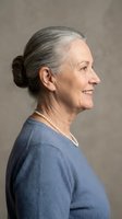 Elegant senior Caucasian woman with gray hair in bun wearing blue sweater and pearl necklace smiling in profile against neutral background.