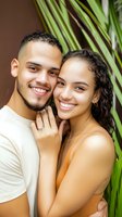 Young Latin American couple smiling and embracing outdoors, showing engagement ring. Natural palm leaves in background create tropical romantic atmosphere.