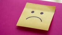 Yellow sticky note with sad face drawn on pink background expressing negative emotions and disappointment in office or workplace setting.