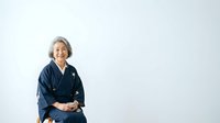 Senior woman with gray hair wearing traditional dark blue kimono, smiling warmly while seated against white background, suitable for cultural, lifestyle, and portrait projects.