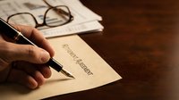 Hand writing last will and testament document with pen on wooden desk next to eyeglasses and papers for estate planning and legal preparation.