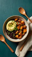 Healthy Buddha bowl with roasted sweet potato cubes, sliced avocado, black beans, and fresh herbs in ceramic dish on teal background. Perfect for food blogs, wellness content, and nutrition articles.