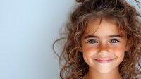 Young girl with bright blue eyes and curly brown hair smiling warmly against light background, expressing joy and innocence in natural lighting portrait.
