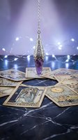 Silver pendulum with purple crystal hanging over tarot cards spread on dark marble surface with fairy lights creating mystical atmosphere for divination.