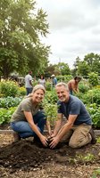 Two middle-aged Caucasian gardeners planting seedlings together in community garden bed, surrounded by growing vegetables and other volunteers working in background. Urban farming and sustainable agriculture concept.