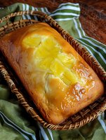 Freshly baked lemon pound cake with glazed citrus slices on top, served in rustic wicker basket. Golden crust glistens with syrup drops on green striped cloth.