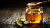 Golden honey dripping from wooden spoon into glass jar with fresh cucumber slices on rustic wooden surface for natural skincare and wellness.