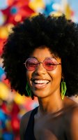 Happy young African American woman with afro hairstyle wearing pink sunglasses and green earrings smiling joyfully against colorful bokeh background.