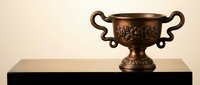 Ornate bronze trophy cup with decorative handles and floral engravings on black pedestal base, isolated on beige background for awards and recognition designs.
