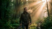 Adult male with beard, wearing dark outdoor jacket, walking through sunlit forest path surrounded by green trees and golden light rays filtering through canopy. Suitable for outdoor adventure, lifestyle, and nature-themed projects.