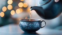 Steaming hot tea being poured from black teapot into ornate ceramic cup with floral pattern against soft bokeh lights background creating cozy atmosphere.