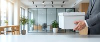 Businessman carrying cardboard box in modern office space during relocation or job transition with glass conference room background.