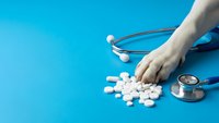 Doctor hand with stethoscope reaching for white pills and tablets on bright blue medical background for healthcare and pharmaceutical concepts.