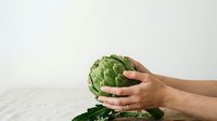 Hands holding fresh green artichoke on white surface for cooking, food preparation, healthy eating, and culinary design projects.