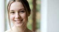 Young Caucasian woman with brown hair smiling warmly by window with natural light creating soft portrait for lifestyle and wellness content.