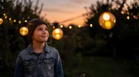 Young woman in denim jacket enjoying warm evening outdoors with glowing string lights creating magical ambiance during golden hour sunset.