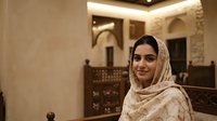 Young Middle Eastern woman wearing traditional beige hijab headscarf smiling warmly in elegant Islamic architecture interior with ornate decorative elements.