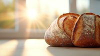 Fresh crusty artisan sourdough bread loaf with golden crust and flour dusting against sunlit window background, morning baking atmosphere.