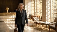 Professional blonde businesswoman in dark suit walking through modern office lobby with geometric sunlight patterns from windows.
