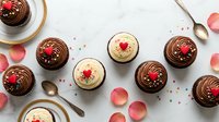 Assorted chocolate and vanilla cupcakes with heart decorations arranged on white marble surface with rose petals and golden spoons for Valentine's Day celebration.
