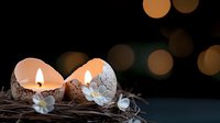 Cracked eggshells with glowing candles inside, decorated with small white flowers and twigs, creating magical Easter or spring atmosphere against dark bokeh background.