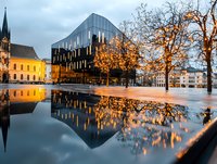 Modern glass building with dramatic reflections in water puddle at dusk, illuminated trees and historic architecture create atmospheric urban scene.