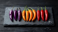 Colorful chili peppers arranged in a gradient from purple to red on a dark slate board with sea salt, top view.