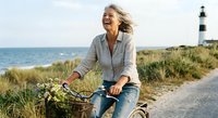 Joyful mature blonde woman cycling near coastal lighthouse on sunny day, expressing freedom and active lifestyle in seaside environment.