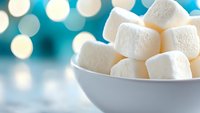 White fluffy marshmallows in ceramic bowl against blurred turquoise background with bokeh lights, creating cozy winter atmosphere for food photography.