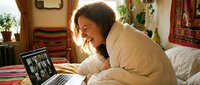 Young Caucasian woman laughing during video call on laptop, wrapped in blanket on bed in cozy bohemian bedroom, remote work and virtual meeting concept.