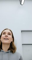 Young woman with shoulder-length brown hair and fair complexion wearing gray hoodie, looking upward with cheerful expression in modern minimalist interior space.