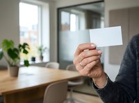 Professional holding blank white card in modern office space with wooden desk, potted plants, and bright windows in background.