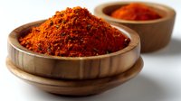 Red chili powder or paprika spice in wooden bowl on white background, macro food photography showing vibrant color and texture of ground spices.