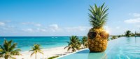 Tropical pineapple with sunglasses by infinity pool overlooking turquoise ocean and palm trees, perfect summer vacation concept for travel and leisure marketing.