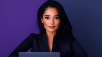 Young Asian businesswoman with long dark hair working on laptop against purple background, professional corporate portrait in black blazer.