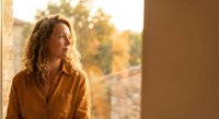 Young woman with curly hair in mustard shirt gazing thoughtfully out window during golden hour sunset, warm natural lighting creating peaceful contemplative mood.