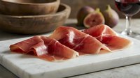 Sliced prosciutto ham on white marble cutting board with fresh figs and wooden bowls in rustic kitchen setting for gourmet cooking and appetizers.