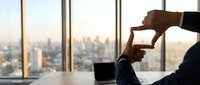 Business professional making hand frame gesture at office window overlooking city skyline during golden hour for creative vision and planning concepts.
