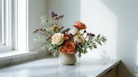 Elegant floral arrangement with peach and orange roses, purple wildflowers, and greenery in a ceramic vase on a marble surface near a window.