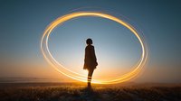 Silhouette of person standing in field at sunset with glowing light trail forming circular halo around them, creating magical atmosphere.