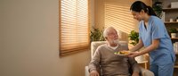 Asian female nurse in blue scrubs serving healthy meal to elderly Caucasian man in comfortable home care setting with warm natural lighting.