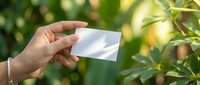 Hand holding a blank white business card against a lush green garden background, mockup with copy space for branding design.