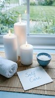 Serene self care spa setup with lit white candles, bath salts, rolled towel, and handwritten note on bamboo mat by window.